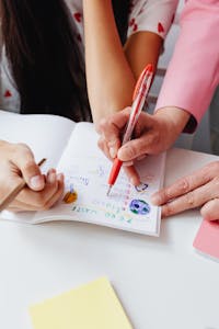 Close-up of hands guiding a student in writing and drawing in a notebook, emphasizing education and creativity.